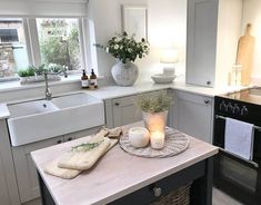 A wooden-countered kitchen island sits in the middle of this small kitchen, decorated with a pair of wooden chopping boards, a wicker tray, a pair of candles, and a plant. Gray cabinets sit next to white countertops and a farmhouse sink...   Image: home_at_ivy_corner