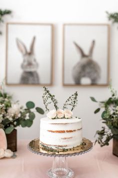 a white cake sitting on top of a table next to two framed pictures and greenery