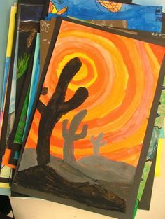 some paintings are on the table and there is a cactus in the foreground with an orange sky behind them