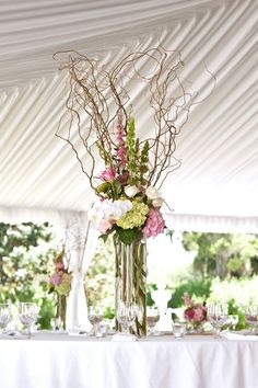 flowers and branches in vases on a table under a white tented area with wine glasses