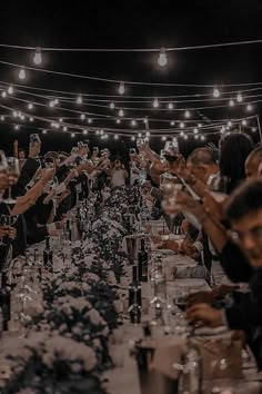 a group of people sitting at a table with wine glasses in their hands and lights strung above them