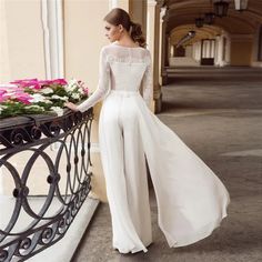 a woman standing on a balcony next to flowers wearing a long white dress and high heels