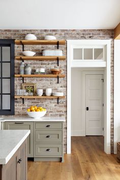 a brick wall in a kitchen with open shelving above the sink and below it is a bowl of fruit