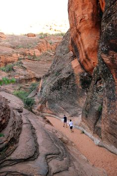 two people are walking along a narrow path in the desert near some large rock formations