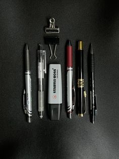 several pens and other items laid out on a black surface with a clipboard in the background