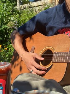 a person sitting down playing an acoustic guitar in front of some plants and flowers with a bottle of ketchup next to them