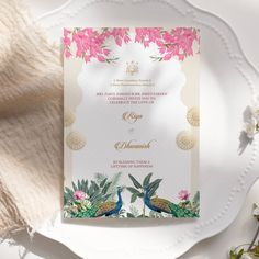 the wedding card is decorated with pink flowers and green leaves, along with two peacocks