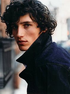 a black and white photo of a young man with curly hair wearing a pea coat