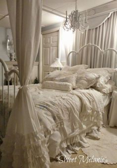 a white bed sitting in a bedroom next to a chandelier and window with curtains