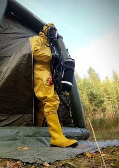 a man in yellow coverall and boots standing next to a black tent with trees in the background