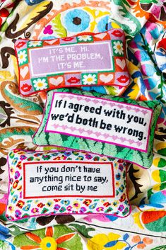 three colorful pillows with embroidered sayings on them sitting on a floral print fabric covered surface