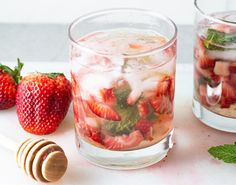 two glasses filled with ice and strawberries on top of a table next to a honey shaker