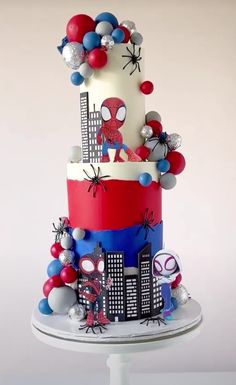 a three tiered cake decorated with balloons and spiderman decorations on a white pedestal