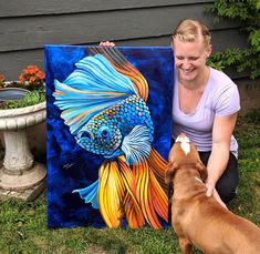 a woman kneeling down next to a painting with a fish on it and a dog looking at it