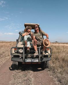 two people sitting in the back of a vehicle on a dirt road with dry grass