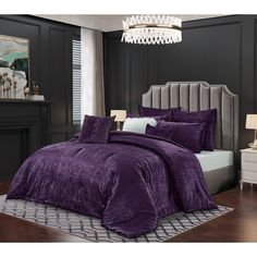 a bed with purple comforter and pillows in a dark colored room, next to a chandelier