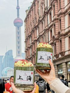 two people holding up jars with food in front of them on a city street near tall buildings