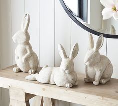 three statues of rabbits sitting on a wooden table in front of a mirror and flower vase