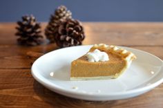 a slice of pumpkin pie on a white plate next to some pine cones and twine needles