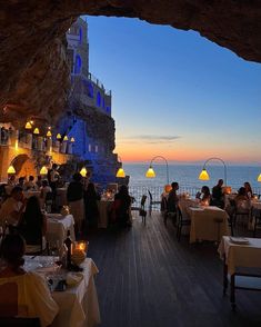 people sitting at tables in front of an ocean view restaurant with lights on the walls