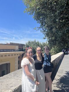 three young women standing next to each other on a sidewalk near trees and buildings in the background