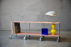 a table with two shelves and a lamp next to each other on wheels in front of a concrete wall