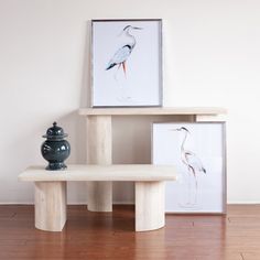 two paintings and a vase on a wooden table in front of a white wall with wood flooring