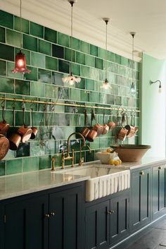 a kitchen with green tiles and copper pots hanging on the wall above the sink, along with brass faucets