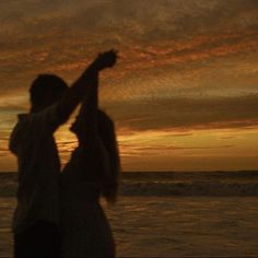 two people are standing on the beach at sunset with their arms up in the air