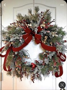 a christmas wreath hanging on the front door with red ribbon and evergreens, berries, pine cones and holly