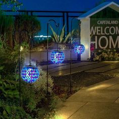 some lights that are on the side of a building near plants and trees in front of a welcome home sign