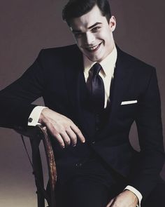 a man in a suit sitting on a chair with his hands on his knees and smiling