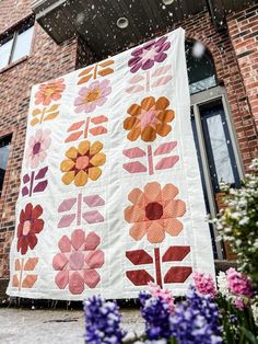 a quilt hanging from the side of a brick building with flowers in front of it