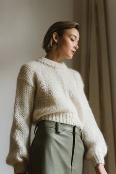 a woman standing in front of a window wearing a white sweater and khaki pants