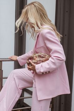 a blonde woman in pink suit and heels sitting on a metal railing holding a purse