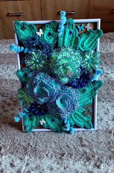 an arrangement of green and blue crocheted flowers in a white box on a table