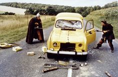 two men standing next to an old car on the side of the road with tools