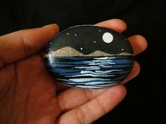 a hand holding a painted rock with mountains and stars in the sky above water at night