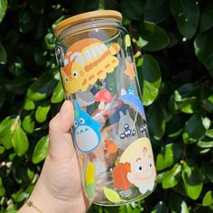 a hand holding a glass with various cartoon stickers on the lid and bottom, in front of some green leaves