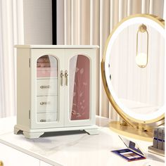 a white cabinet sitting on top of a table next to a mirror and lamp with pink curtains