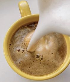 milk being poured into a yellow cup filled with liquid and coffee beans on a white surface