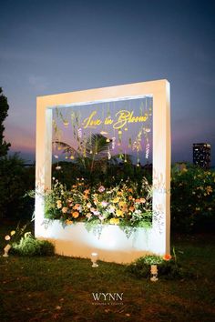 an illuminated sign with flowers on it in the grass at night, surrounded by greenery