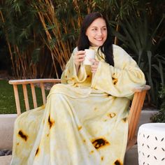 a woman sitting on a wooden bench holding a coffee cup in her hand and wrapped in a blanket