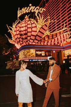 a man and woman holding hands in front of a building with lights on the side