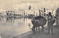 an old photo of two people and a donkey by the water in front of some buildings