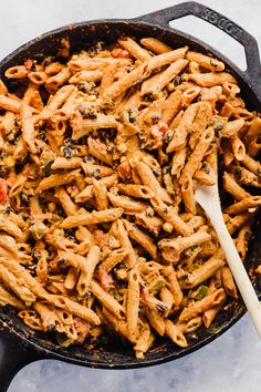 a skillet filled with pasta and vegetables on top of a table next to a wooden spoon