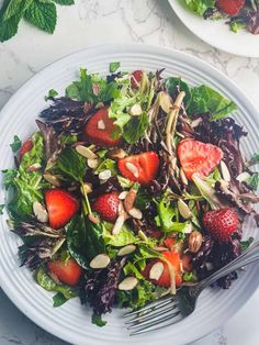 a salad with strawberries, almonds and greens on a white plate next to a fork