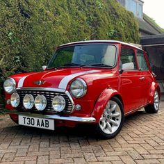 an old red and white mini car parked in front of a house with bushes behind it