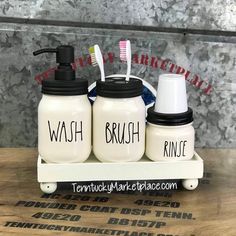 three white jars with toothbrushes in them sitting on a wooden table next to a black and white toothbrush dispenser