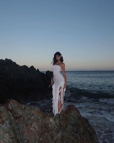 a woman standing on top of a rock next to the ocean at sunset with her dress blowing in the wind